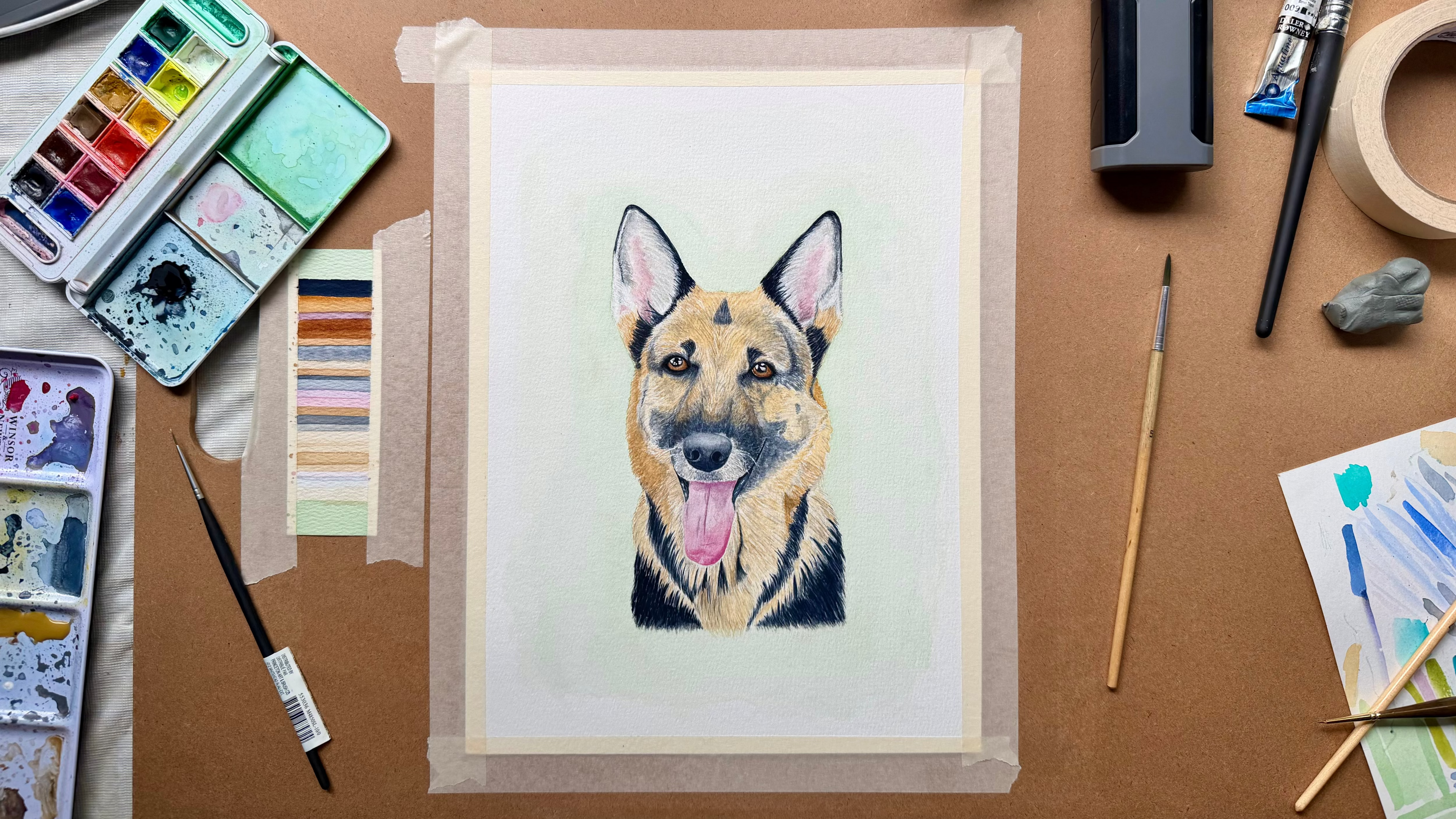 A watercolor painting of a German Shepherd dog on a sheet of paper taped to a work surface, surrounded by art supplies including watercolor palettes and brushes.